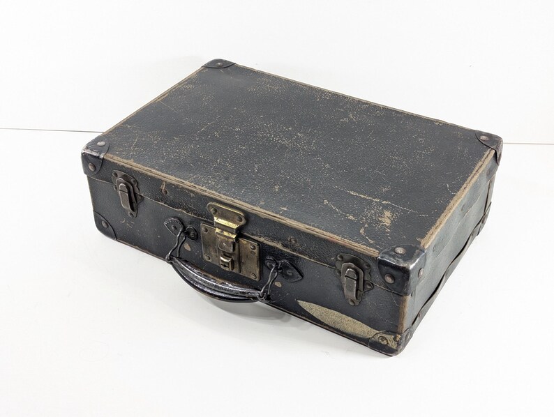 May include: A rectangular black suitcase with a weathered surface, featuring reinforced metal corners, a central brass-toned latch, and two side locking mechanisms. The case includes a black handle attached to the front with metal hardware.