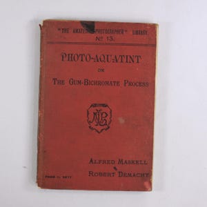 May include: A red book titled "Photo-Aquatint or The Gum-Bichromate Process" by Alfred Maskell and Robert Demachy. The book is part of "The Amateur Photographer Library" series, number 13.