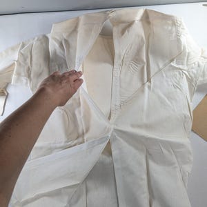 May include: An off-white, long-sleeved shirt with a collared neckline. The shirt is made of a vertically striped fabric. A hand is partially visible, holding open the front of the shirt. The shirt is laid out on a white surface.