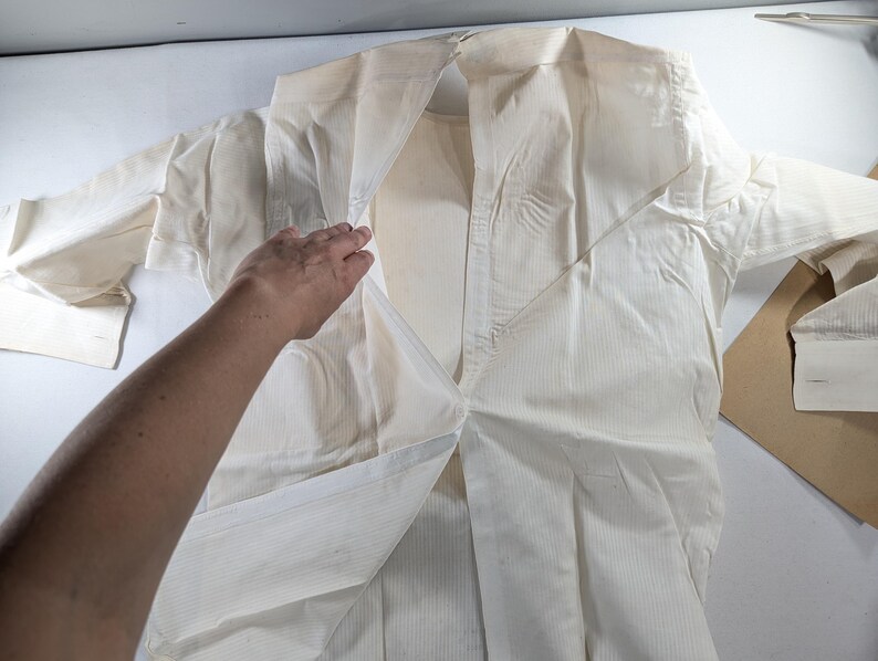 May include: An off-white, long-sleeved shirt with a collared neckline. The shirt is made of a vertically striped fabric. A hand is partially visible, holding open the front of the shirt. The shirt is laid out on a white surface.