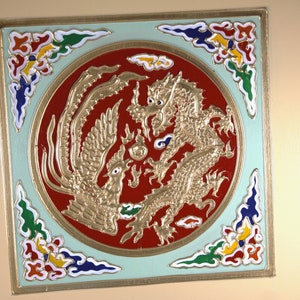 May include: A square, framed artwork with a circular design featuring a gold dragon and phoenix on a red background. The artwork is surrounded by a light green border with colorful floral designs in the corners.