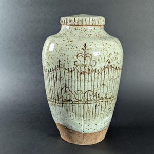 Może przedstawiać: Ceramiczny wazon z jasnozielonym szkliwem i brązowymi plamkami. Wazon posiada ręcznie rysowany wzór ogrodzenia w kolorze brązowym. Podstawa jest naturalna, nieszkliwiona brązowa. Wazon ma zaokrąglony korpus i wąską szyję.