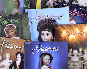 Pode incluir: Uma coleção de revistas de bonecas vintage intituladas "Gildebrief" é exibida. As capas apresentam imagens de bonecas antigas, retratos e temas de férias. As revistas são em várias cores, incluindo verde, azul e castanho.