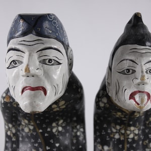 May include: Two wooden figurines of people with painted faces and black and white patterned clothing. The figurines are carved in a traditional style and have a whimsical expression.