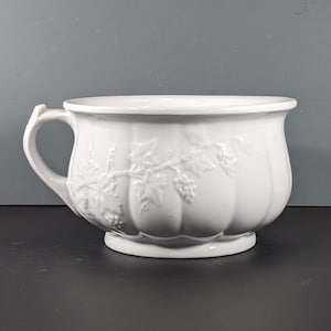 Könnte beinhalten: Weißer Keramiknapf mit Henkel und geprägtem Weinreben-Design. Die Schüssel hat eine weite Öffnung und einen abgerundeten Boden. Der Griff ist gebogen und an der Seite befestigt. Ideal zum Servieren oder Präsentieren.