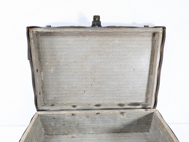 May include: An open rectangular trunk with a dark brown exterior and a light gray fabric-lined interior. The trunk features a wooden frame structure, a metal latch mechanism at the top center, and visible metal rivets along the inner edges.