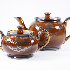 Könnte beinhalten: Ein braunes Keramik-Teekannen- und Zuckergefäß-Set mit silbernen Blumendesigns. Die Teekanne hat einen Deckel und einen Griff. Das Zuckergefäß hat einen Deckel und einen Griff.