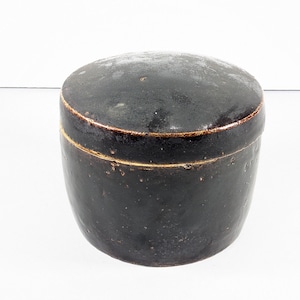 May include: A dark brown, lidded ceramic container with a rounded shape. The lid and body are a deep, mottled brown with a slightly glossy finish. A thin, gold-colored band accents the seam between the lid and the body.