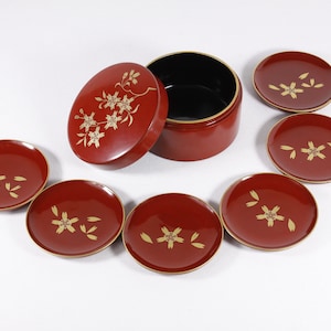 May include: A set of seven red lacquer coasters with gold floral designs. The coasters are arranged in a circle around a red lacquer box with a black interior. The box has a lid with a matching floral design.
