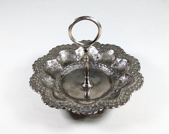 May include: A silver-toned metal serving tray with a scalloped edge and a central vertical handle featuring a circular ring at the top. The tray surface is decorated with an intricate embossed floral pattern throughout the scalloped rim area.