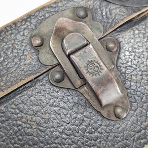 May include: A black textured case featuring a weathered metal latch mechanism. The latch is secured with four rivets and displays a small circular emblem with a sunburst design and the letter P in the center, showing signs of age and oxidation.