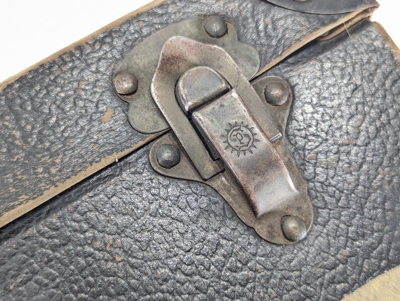 May include: A black textured case featuring a weathered metal latch mechanism. The latch is secured with four rivets and displays a small circular emblem with a sunburst design and the letter P in the center, showing signs of age and oxidation.