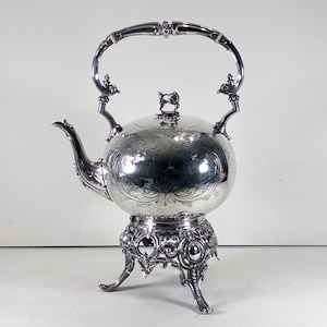 May include: A polished silver kettle with an ornate, engraved floral pattern on its spherical body. It features a curved spout, a high arched handle, and a lid with a small finial. The kettle rests on a decorative, openwork metal stand with three legs.
