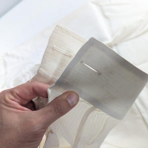 May include: Close-up of a cream-colored shirt cuff with a buttonhole. The cuff is held up, showing the fabric's texture and the buttonhole's detail. The shirt appears to be made of a lightweight material.