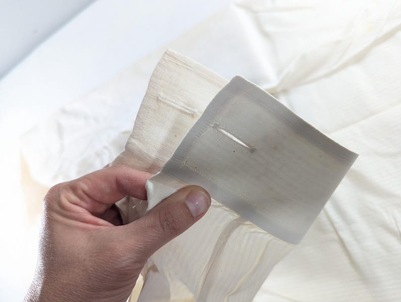 May include: Close-up of a cream-colored shirt cuff with a buttonhole. The cuff is held up, showing the fabric's texture and the buttonhole's detail. The shirt appears to be made of a lightweight material.