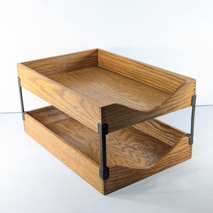 Könnte beinhalten: Ein zweistufiger Schreibtisch-Organizer aus Holz mit natürlicher Holzmaserung. Die Ablagen werden von schwarzen Metallwinkeln getragen. Das obere Fach hat eine gebogene Vorderkante. Für Büro oder Zuhause.