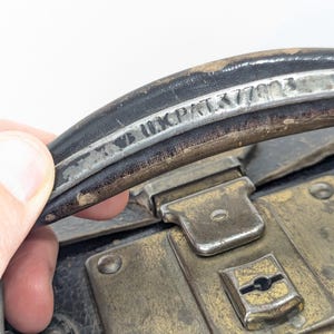 May include: A black leather suitcase handle featuring a metallic strip with the embossed text "UK PAT 377402". The handle is attached to a black suitcase body with a tarnished brass-colored metal latch mechanism featuring a central keyhole.