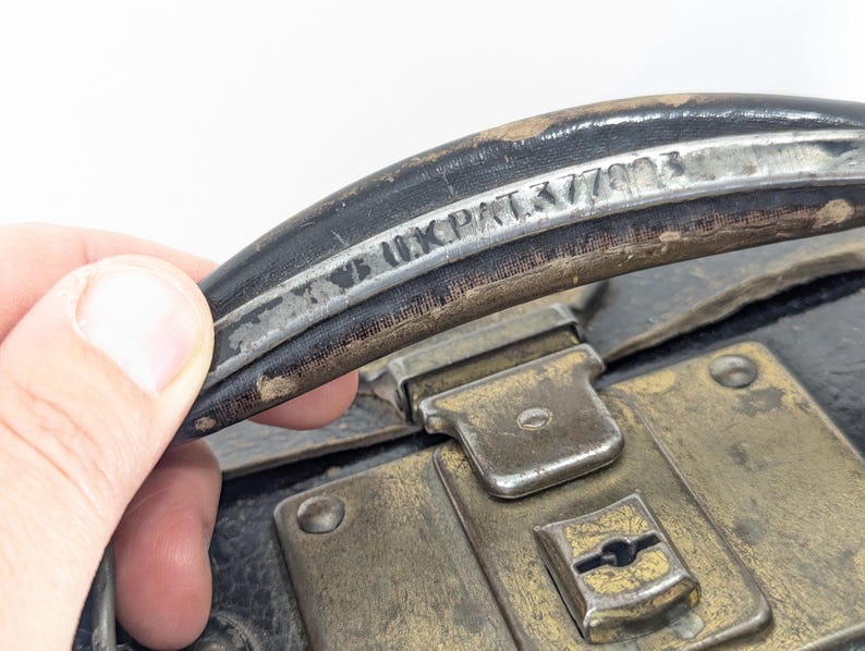 May include: A black leather suitcase handle featuring a metallic strip with the embossed text "UK PAT 377402". The handle is attached to a black suitcase body with a tarnished brass-colored metal latch mechanism featuring a central keyhole.