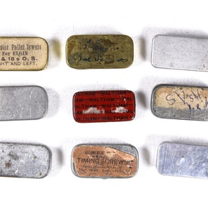 May include: Nine small metal tins with various labels. The labels include "Excelsior Pallet Jewels For ELGIN 16 & 18 s O. S. RIGHT AND LEFT.", "Waltham", "Assbrement Timing Sorews", and "S.V.S.H."