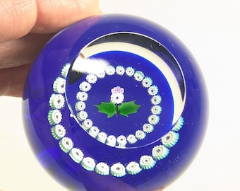 May include: A spherical cobalt blue glass paperweight featuring a central millefiori design of a white and purple thistle with green leaves, surrounded by two concentric circular rings of small white and light blue floral canes.