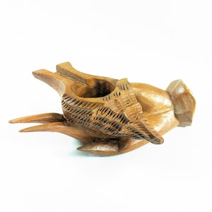 May include: A hand-carved wooden sculpture of a bird holding a basket. The bird's wings and the basket are intricately detailed. The sculpture is made of light brown wood and sits on a white background.