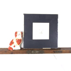 May include: A vintage wooden level with a ceramic dog figurine and a dark blue book with a white square on a white background. The dog is white with red spots.