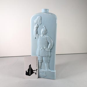 Puede incluir: Una botella de cerámica azul claro con un relieve en relieve de una figura de guerrero. La figura sostiene una pancarta. Una tarjeta blanca con un logotipo de cuervo negro y texto está frente a la botella.
