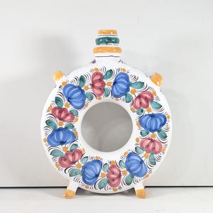 May include: A white ceramic ring-shaped vessel with a central circular opening, featuring hand-painted floral motifs in blue, red, and green. The neck has yellow and green bands, and the base includes two small yellow feet for stability.