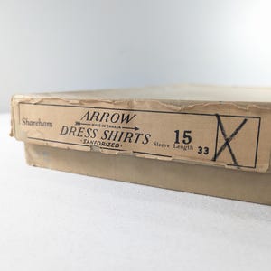 May include: A vintage cardboard box with a label that reads "ARROW DRESS SHIRTS." The label also indicates the shirt size is 15 and the sleeve length is 33 inches. The box is tan and has a worn appearance.