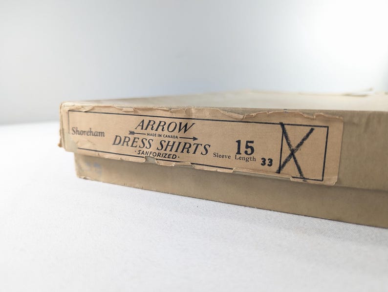 May include: A vintage cardboard box with a label that reads "ARROW DRESS SHIRTS." The label also indicates the shirt size is 15 and the sleeve length is 33 inches. The box is tan and has a worn appearance.