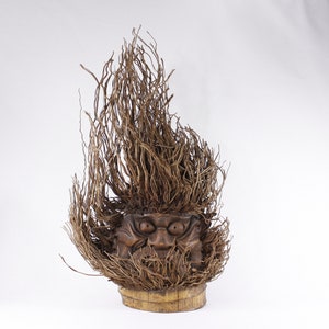 Könnte beinhalten: Eine Holzskulptur eines Gesichts mit wildem, buschigem Haar aus Naturfasern. Das Gesicht hat einen geschnitzten Ausdruck von Wut oder Überraschung. Die Skulptur ist auf einem Holzsockel montiert.