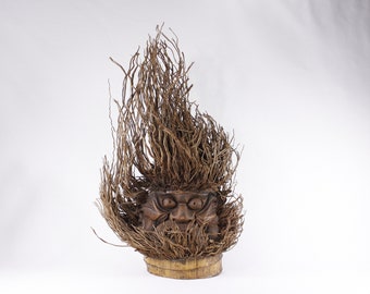 May include: A wooden sculpture of a face with a wild, bushy hair made of natural fibers. The face has a carved expression of anger or surprise. The sculpture is mounted on a wooden base.