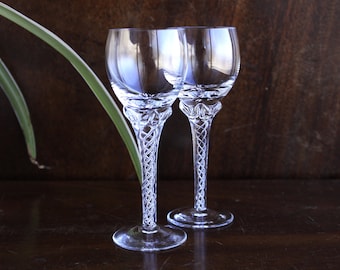 May include: Two clear glass wine glasses with a twisted stem design. The glasses are empty and sitting on a dark wooden surface.