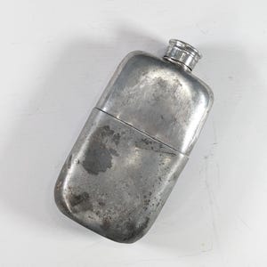 Puede incluir: Una petaca metálica rectangular antigua con tapón de rosca. La petaca tiene un aspecto desgastado y envejecido, con signos visibles de uso. El metal es de color gris plateado opaco con manchas oscuras. La petaca está vacía.