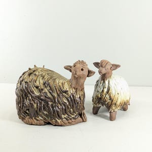 Puede incluir: Dos figuras de ovejas de cerámica esculpidas a mano. La oveja más grande está en posición reclinada con lana texturizada en marrón oscuro y bronceado. La oveja más pequeña de pie presenta un pelaje esmaltado en blanco y marrón con detalles.