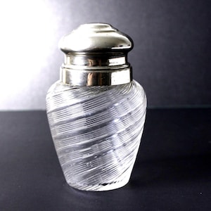 May include: A clear glass jar featuring a distinct diagonal ribbed texture throughout its body. The jar is topped with a smooth, polished silver-toned metal lid that fits securely over the neck, creating a contrast between the glass and metal.