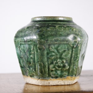 Peut inclure: Un pot en céramique vert de forme hexagonale avec des motifs floraux complexes. Le pot a une texture légèrement rugueuse et une base brun clair.