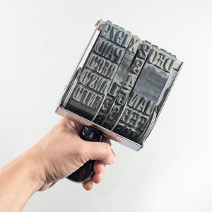 May include: A vintage metal date stamp with a black handle. The stamp features multiple rotating bands with letters and numbers. The stamp is held against a white background.