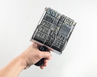 May include: A vintage metal date stamp with a black handle. The stamp features multiple rotating bands with letters and numbers. The stamp is held against a white background.