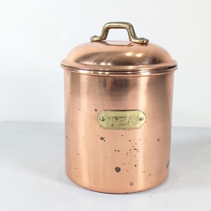 May include: A copper tea canister with a lid and a brass handle. The canister has a brass label that reads "TEA". The copper has a warm, reddish-brown hue and a slightly aged appearance.
