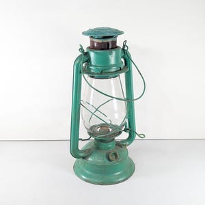 May include: Vintage teal-colored lantern with a clear glass globe. The lantern has a metal handle and a round base. The top of the lantern is a darker teal color. The lantern is designed to be used with oil or kerosene.