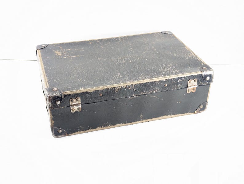 May include: A rectangular black storage box with a worn, distressed finish. The box features silver-toned metal corner protectors and two silver-toned metal hinges along the front edge. The surface shows visible signs of age and wear throughout.