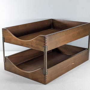 May include: A two-tier wooden document tray featuring dark brown wood construction with visible finger joints at the corners. The trays are connected by vertical metal brackets on each side, and each tray has a curved cutout on the front panel.