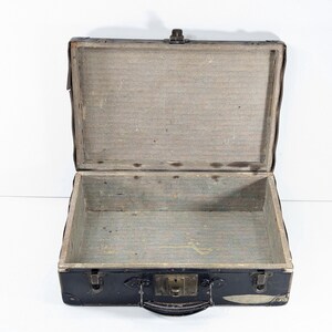 May include: An open vintage suitcase with a black exterior and a light gray interior lining. The rectangular case features a central metal latch, a top handle, and reinforced corners. The interior is empty, showing a plain, textured surface.