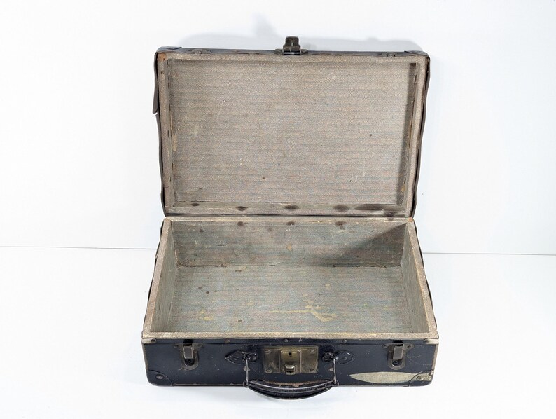 May include: An open vintage suitcase with a black exterior and a light gray interior lining. The rectangular case features a central metal latch, a top handle, and reinforced corners. The interior is empty, showing a plain, textured surface.