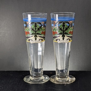 May include: Two tall, clear glass beer mugs with blue and yellow trim. The mugs have a colorful design featuring a tree and the word "Madeira" on the front.