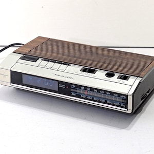 May include: A Realistic brand digital clock radio with a wood-grain top panel and a silver-toned front face. The device features a digital time display, an analogue AM/FM radio tuner with a red needle, volume and tuning dials, and top-mounted buttons.