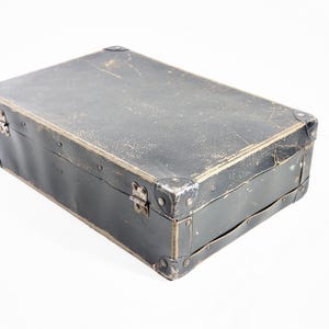May include: A rectangular black suitcase with a weathered, textured surface. The case features reinforced metal corner protectors, silver-toned metal hinges, and a side handle. The exterior shows signs of wear with visible scuffs and scratches.