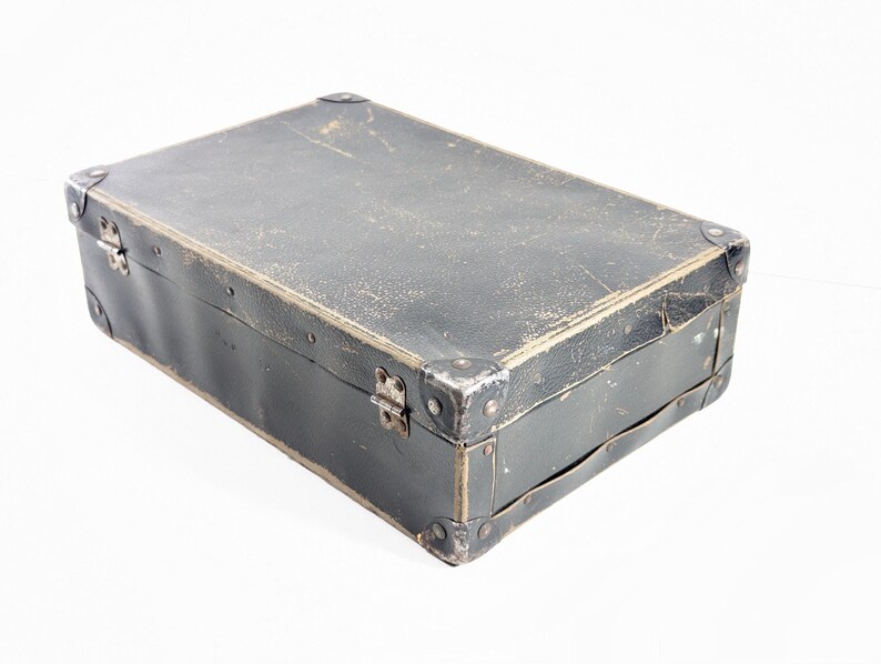 May include: A rectangular black suitcase with a weathered, textured surface. The case features reinforced metal corner protectors, silver-toned metal hinges, and a side handle. The exterior shows signs of wear with visible scuffs and scratches.