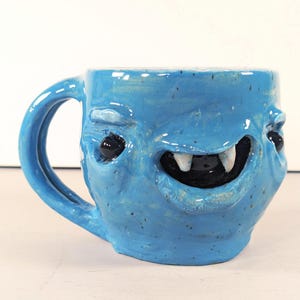 Puede incluir: Una taza de cerámica hecha a mano con un diseño de cara de monstruo caprichoso. La taza está pintada en un color azul vibrante, con ojos negros, una amplia sonrisa con colmillos blancos y un asa azul a juego. La taza es una obra de arte única.